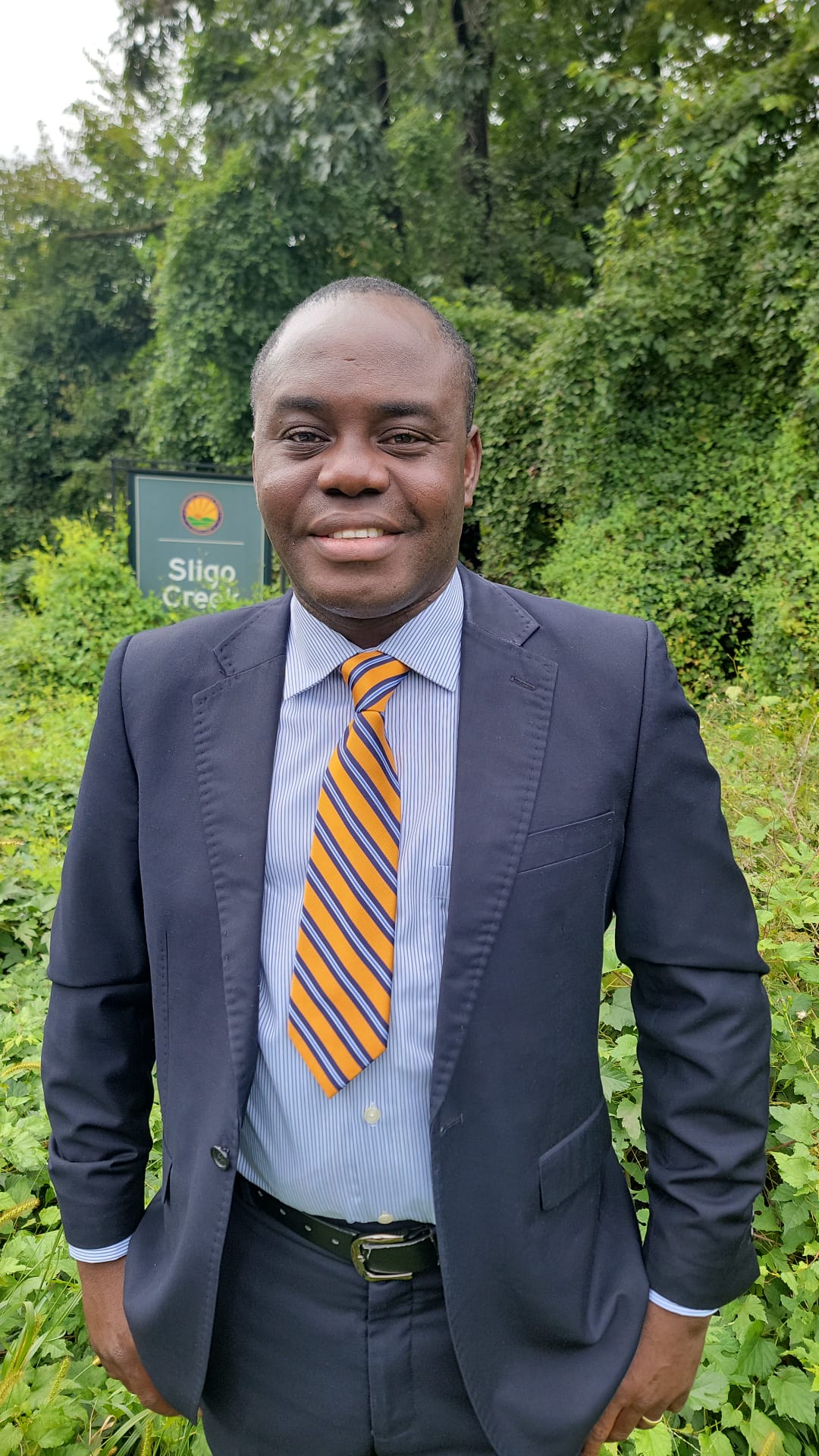 Ambroise Agosse standing outdoors near Sligo Creek, wearing a navy suit and orange striped tie